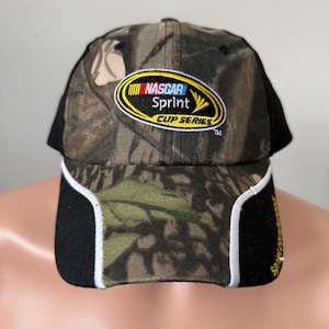 Puede incluir: Una gorra de béisbol de camuflaje negro y verde con un parche ovalado amarillo que dice "NASCAR Sprint Cup Series" en texto negro.