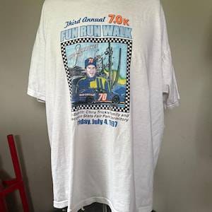 May include: White t-shirt featuring a colorful graphic for the Third Annual 7.0K Fun Run Walk. The graphic includes a race car, a driver, and the text "Milwaukee The Mile". The shirt also has text that reads "Friday, July 4, 1997".