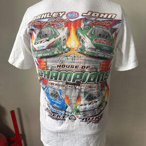 May include: White t-shirt featuring a colorful graphic of race cars and the text "ASHLEY JOHN FORCE" and "HOUSE OF CHAMPIONS". The design includes the NHRA logo and various sponsor logos. The shirt has short sleeves and a crew neck.