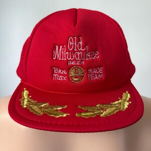 May include: Red mesh trucker hat with a white embroidered logo that says "Old Milwaukee Beer" with a gold circle and the words "Blue Max Race Team". The hat has gold embroidered feathers on the brim.