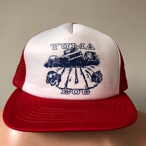 Puede incluir: Una gorra de camionero roja y blanca con un gráfico azul de vehículos todoterreno y el texto "YUMA BOG 1986".