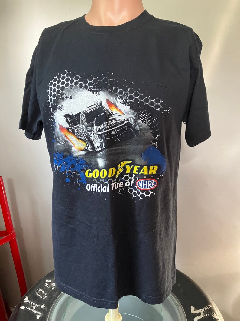 Pode incluir: Camiseta preta com um gr&aacute;fico de um carro de corrida com chamas e o texto "Goodyear Official Tire of NHRA".