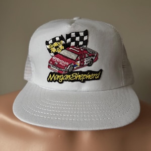 May include: White trucker hat with a curved brim and mesh back. The front features an embroidered patch with a red race car, checkered flags, and the text "Tested Tough" and "Morgan Shepherd".