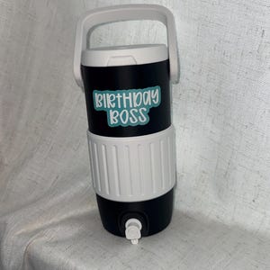 Puede incluir: Nevera aislada negra y blanca con asa blanca. La nevera tiene una banda blanca en el medio y las palabras "BIRTHDAY BOSS" en verde azulado. Un grifo es visible en la parte inferior.