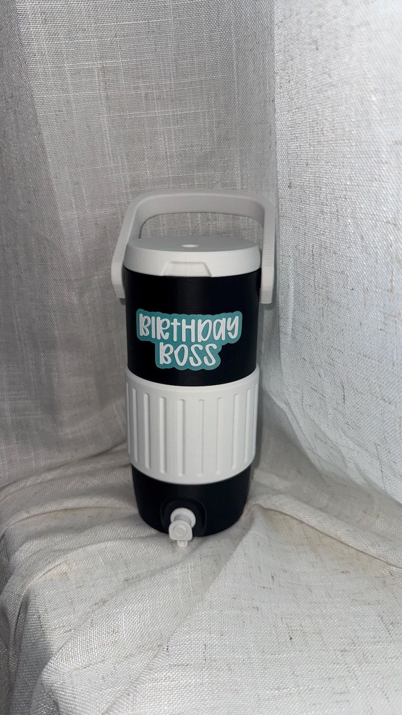 Puede incluir: Enfriador de bebidas aislado en blanco y negro con asa y tapa blancas. El enfriador tiene el texto "BIRTHDAY BOSS" en azul claro. Tiene un grifo para dispensar bebidas.
