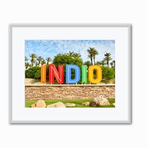 May include: Framed photograph featuring the word "INDIO" in large, colorful letters. The letters are set against a backdrop of palm trees, a blue sky with clouds, and a stone wall. The image is framed in white.