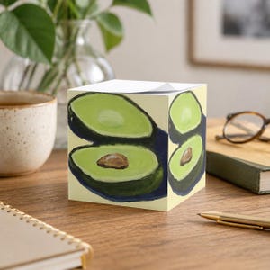 Puede incluir: Un bloc de notas cúbico con un diseño de aguacate pintado. La obra de arte representa aguacates cortados por la mitad con pulpa verde, contornos verde oscuro y huesos marrones, sobre un fondo amarillo claro. El bloc de notas está sobre una superficie de madera.