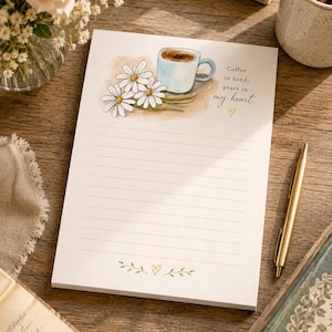Pode incluir: Uma folha de papel de carta pautada branca com uma ilustração em aguarela de três margaridas brancas e uma caneca de café azul-clara no topo. O texto diz "Coffee in hand, peace in my heart." com um pequeno ícone de coração abaixo.
