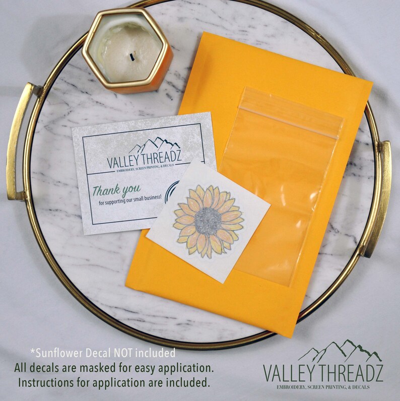 May include: An overhead shot of a package containing a sunflower decal, a thank you card, and a candle on a decorative tray. The package is yellow, and the decal is a watercolor-style image of a sunflower. The text on the card reads "Thank you for supporting our small business!"
