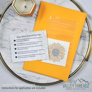 May include: An orange envelope containing a sunflower decal and application instructions. The decal is in a clear plastic bag. The instructions detail how to apply the decal. A gold tray and candle are in the background.
