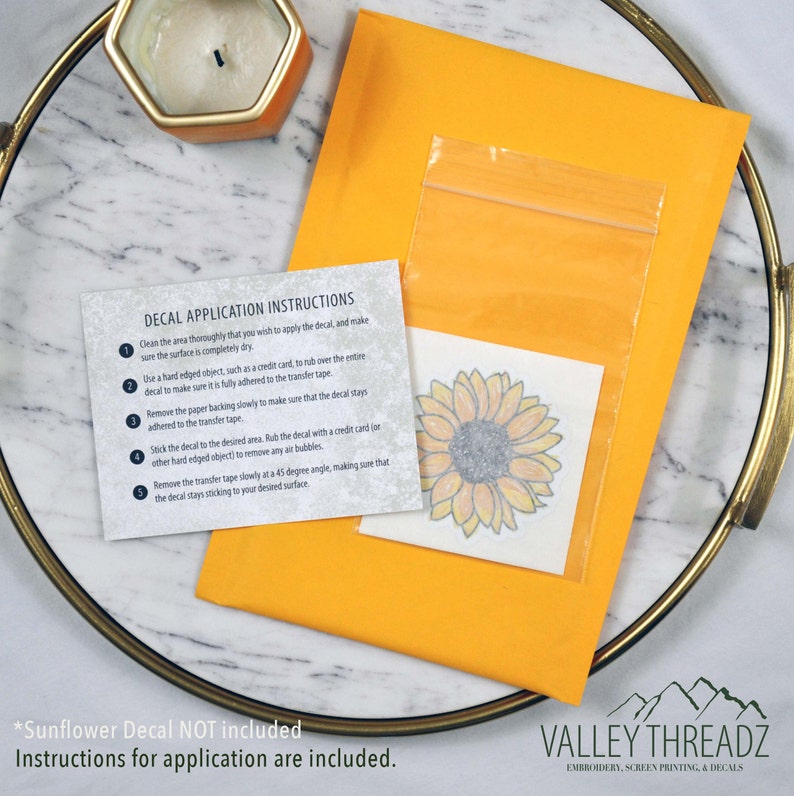 May include: An orange envelope containing a sunflower decal and application instructions. The decal is in a clear plastic bag. The instructions detail how to apply the decal. A gold tray and candle are in the background.