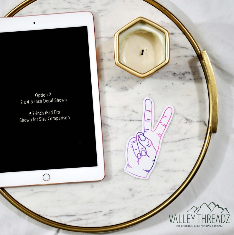 May include: A white sticker with a hand making a peace sign, with a gradient of purple to pink. The sticker is on a marble tray with a gold rim, next to a gold hexagonal candle holder and an iPad. Text on the iPad reads: Option 2, 2 x 4.5-inch Decal Shown, 9.7-inch iPad Pro Shown for Size Comparison.