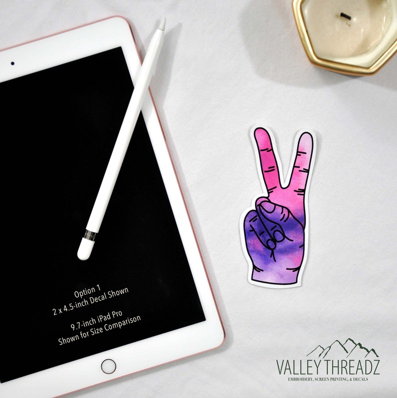 May include: A watercolor-style sticker of a hand making the peace sign, with pink and purple hues. The sticker is next to a 9.7-inch iPad Pro and a white stylus. The text on the iPad reads: Option 1, 2 x 4.5-inch Decal Shown, Shown for Size Comparison.
