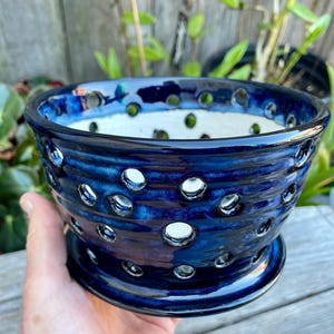 May include: A handcrafted, dark blue ceramic orchid pot with a glossy finish. The pot features numerous circular openings around the sides and a matching saucer base. The interior is white, providing a striking contrast to the exterior. Ideal for orchids or other plants.