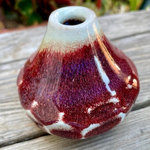 May include: A small, handcrafted ceramic vase with a unique shape. The vase features a deep burgundy glaze that transitions to a lighter, crackled white and blue at the top. The vase has a bulbous body and a narrow opening.