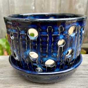 May include: A cylindrical ceramic planter with a deep blue and black glossy glaze, featuring a series of circular cutouts arranged in vertical columns around the body, accompanied by a matching circular saucer base for drainage.