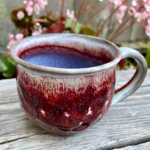 May include: A handmade ceramic mug with a unique glaze. The mug features a deep red exterior that fades to a lighter color near the rim, with a blue interior. The handle is a light gray color. This mug is perfect for coffee or tea.