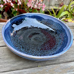 May include: A handmade ceramic bowl with a deep, rounded shape. The bowl has a glossy, dark blue interior with speckled accents and a lighter blue rim. The exterior has a textured, glazed finish.