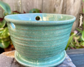 Jardinera colgante de cerámica de color aguamarina. Tiene platillo adjunto, con orificios para drenaje y para colgar. Bonita incorporación para el hogar y el patio.