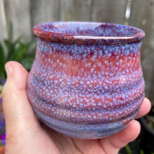 May include: A small, rounded ceramic cup featuring a reactive glaze with a blend of deep red and vibrant blue tones. The surface displays a speckled, mottled texture with horizontal ridges along the body and a smooth, slightly flared rim.