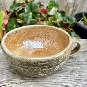May include: A ceramic mug with a wide, rounded bowl shape and a single handle. The exterior features a textured, mottled olive-green glaze, while the interior displays a smooth, glossy, warm honey-orange finish with subtle speckling throughout.