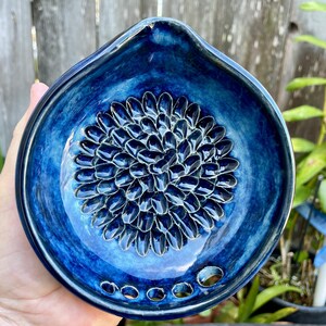 May include: A teardrop-shaped ceramic grater dish featuring a deep blue glaze with lighter blue highlights. The center displays a raised, textured floral pattern with petal-like ridges for grating, and the bottom edge has five small circular holes.