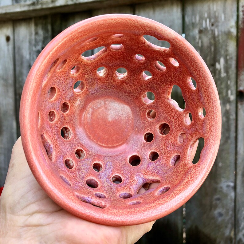 Puede incluir: Un cuenco de cer&aacute;mica de color terracota con un dise&ntilde;o &uacute;nico que presenta un gran agujero central y agujeros m&aacute;s peque&ntilde;os alrededor del borde. El cuenco tiene un esmalte moteado y una superficie ligeramente texturizada.