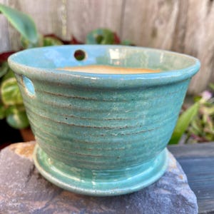 May include: A teal green ceramic planter with drainage holes and a matching saucer. The planter has a textured, striped design.