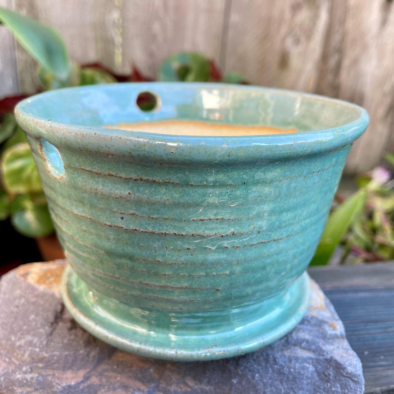 May include: A teal green ceramic planter with drainage holes and a matching saucer. The planter has a textured, striped design.