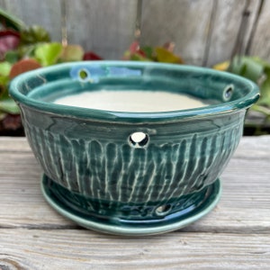 May include: A teal green ceramic planter with drainage holes and a matching saucer. The planter has a textured, vertical pattern.
