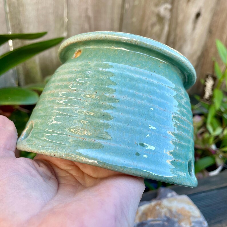 May include: A teal green ceramic planter with a textured, ribbed surface. The planter has drainage holes on the bottom.