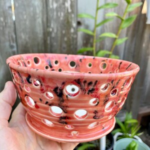 May include: A round ceramic bowl featuring a glossy coral-pink glaze with black mottled accents. The bowl has a wide, flared rim and a series of circular cutouts distributed evenly across its surface, revealing the interior of the vessel.