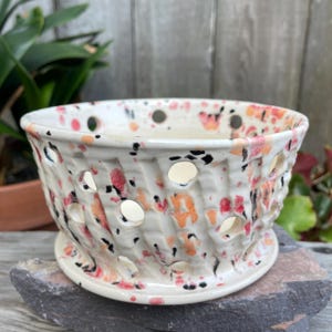 May include: A ceramic bowl with a basket-weave texture and circular cutouts throughout the body. The surface features a speckled glaze with splashes of pink, orange, and black on a cream-colored base, complete with a matching circular base.