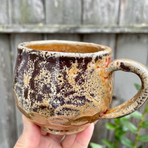 May include: A handmade ceramic mug with a rounded shape and a thick handle. The exterior features a textured, crackled glaze with a colour palette of deep dark brown, warm tan, and subtle orange hues, creating a mottled appearance.