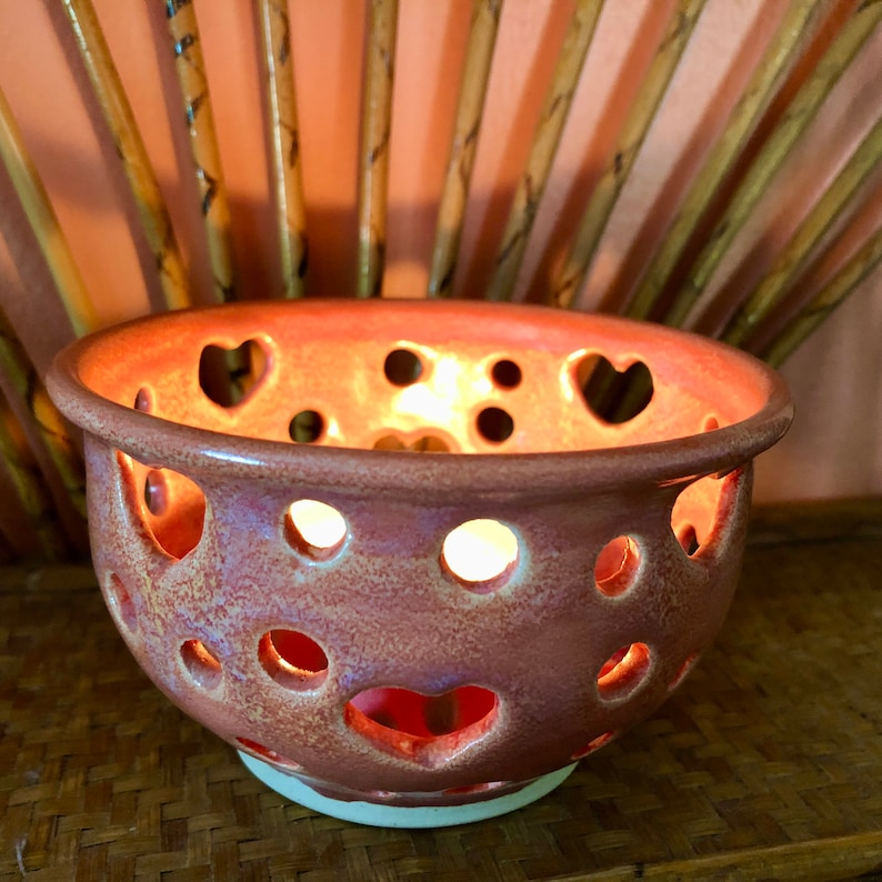 Puede incluir: Un cuenco de cer&aacute;mica de color terracota con recortes en forma de coraz&oacute;n. El cuenco est&aacute; iluminado desde el interior, proyectando un brillo c&aacute;lido.
