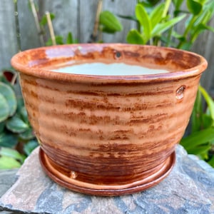 May include: A handcrafted, round ceramic planter with a rich, earthy brown glaze. The planter features a textured, horizontal striped pattern and a matching saucer. Two drainage holes are visible on the side. The interior is a creamy white color.