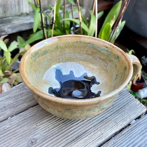 May include: A ceramic bowl with a brown, green, and white glaze. The bowl has a single handle and a black glaze inside.