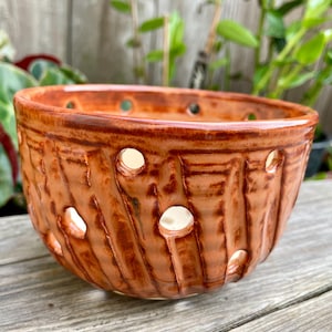 May include: A handmade, terracotta-colored ceramic bowl with a textured surface and decorative cut-out holes. The bowl has a round shape and is suitable for plants or decorative purposes. The bowl is sitting on a wooden surface.