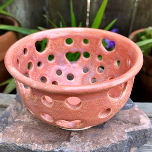 May include: A pink ceramic bowl with a speckled glaze and heart-shaped cutouts. The bowl has a white interior and is perfect for serving fruit or snacks.