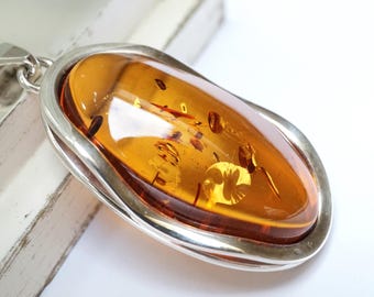 May include: A large, oval amber pendant with a silver-colored bezel. The amber is a warm, translucent orange-brown color, with visible inclusions. The pendant has a simple, elegant design, suitable for everyday wear.