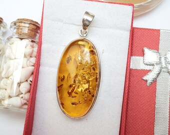 May include: A silver pendant with a large oval amber stone. The amber is a warm honey color and has inclusions of insects and plant matter. The pendant is in a red gift box with a silver ribbon.