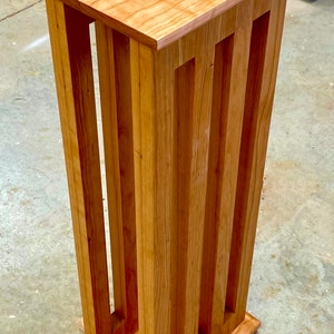 May include: A tall, wooden pedestal stand with a rectangular top and base. The stand's sides feature vertical, evenly spaced cutouts. The wood has a light brown color and visible grain, suggesting a natural finish. The stand is approximately 30 inches tall.