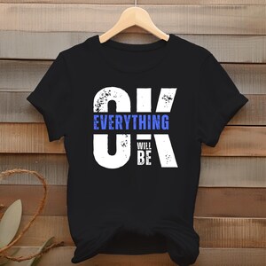 Può includere: T-shirt nera con la scritta "OK Everything Will Be" in bianco e blu. La maglietta è appesa a una gruccia di legno su uno sfondo di legno. La maglietta è un design digitale.