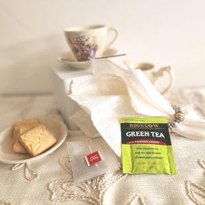 May include: A still life arrangement featuring a teacup and saucer with floral designs, a small plate with cookies, and a Bigelow Green Tea packet. A white napkin and a tea bag are also present, creating a cozy tea time scene.
