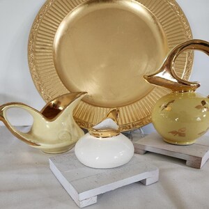 May include: Three vintage ceramic creamer pitchers with gold accents. The pitchers are on white wooden stands. The largest pitcher is yellow with a gold butterfly design. The smallest pitcher is white with a gold rim. The middle pitcher is yellow with a gold rim.