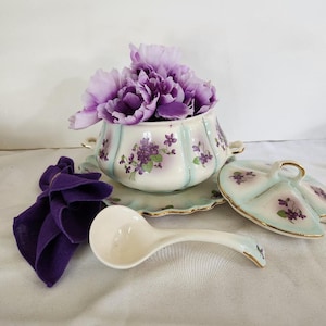 May include: A white porcelain soup tureen with a purple and green floral design. The tureen has a lid and a matching serving spoon. The tureen is filled with purple flowers.