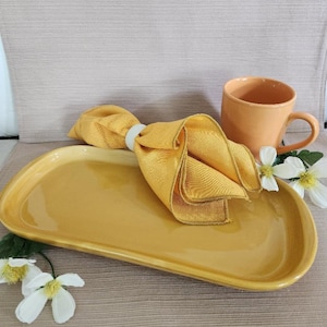 May include: A yellow oval ceramic serving tray with a yellow linen napkin folded on top. The tray is on a brown textured surface.