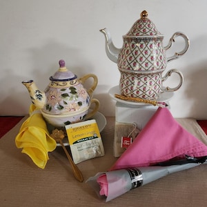 May include: Two teapots, one with a floral design and the other with a pink and white geometric pattern, are displayed on a table with a yellow napkin, a pink napkin, a tea bag, and a tea infuser.  The tea bag is labeled "Bigelow Lemon Ginger Herbal Tea."