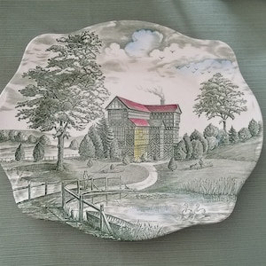 May include: A vintage ceramic plate with a black and white illustration of a Tudor-style house with a red roof, surrounded by trees and a pond with swans. The plate has a scalloped edge and a green background.