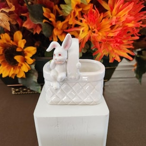 May include: White ceramic bunny figurine sitting in a white ceramic basket with a diamond pattern. The basket is on a white wooden pedestal.